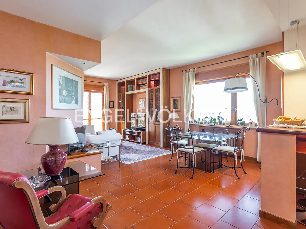 A bright living room with terracotta tile floors, a glass dining table, and a red armchair. A built-in bookcase and large windows add character.