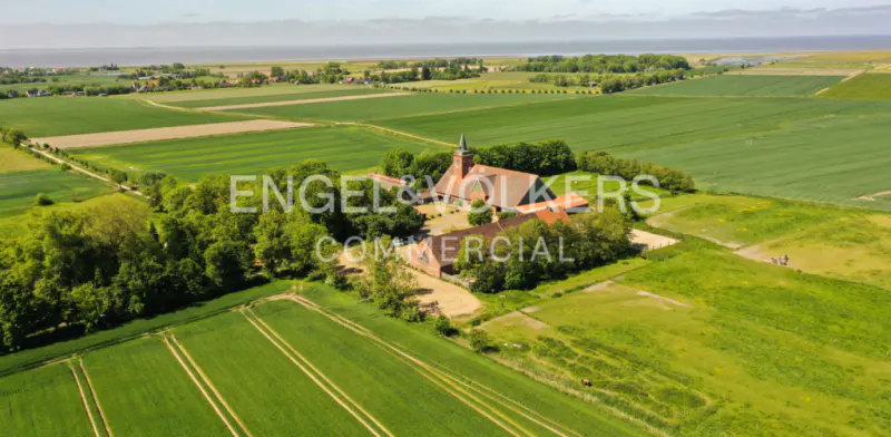 Aerial view of a red brick building with a steeple, surrounded by green fields and trees, near the coast.