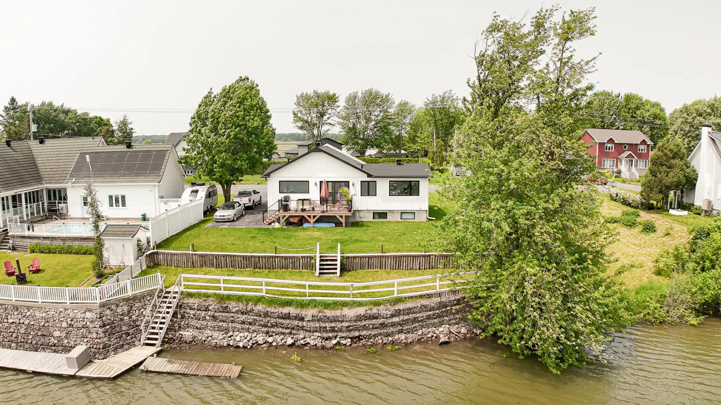 A white house with a black roof sits on a grassy lawn near a body of water. A white fence and stone wall separate the lawn from the water.