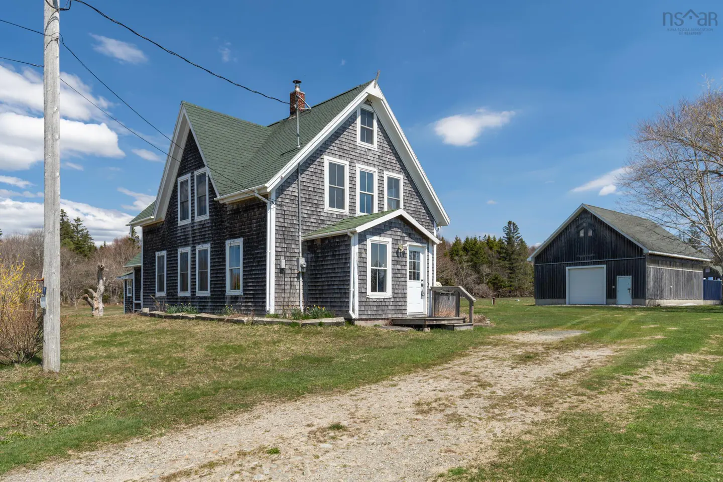 Exterior view of a grey shingle house with a green roof and a detached garage on a sunny day.