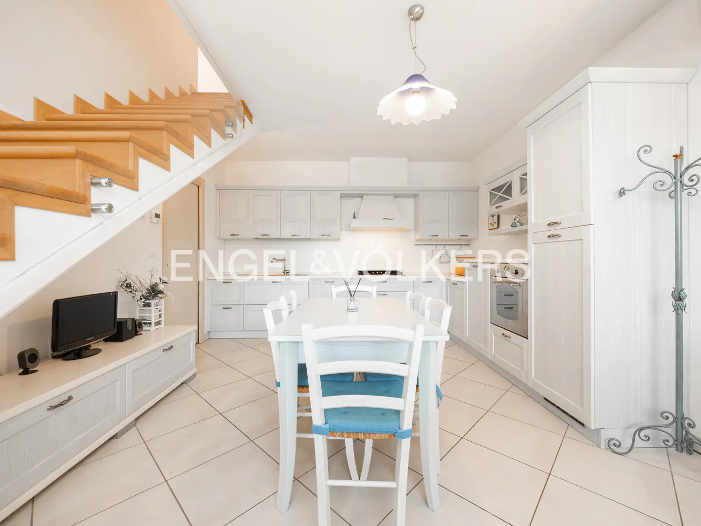 Bright kitchen with white cabinets, a dining table with blue cushions, and a wooden staircase on the left.