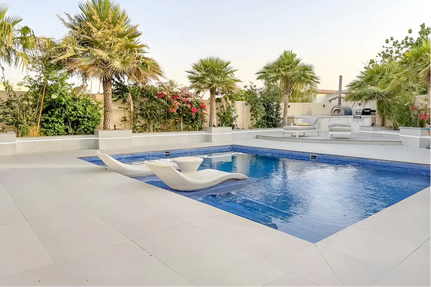 A backyard pool with two white lounge chairs in the water, palm trees, and an outdoor kitchen.