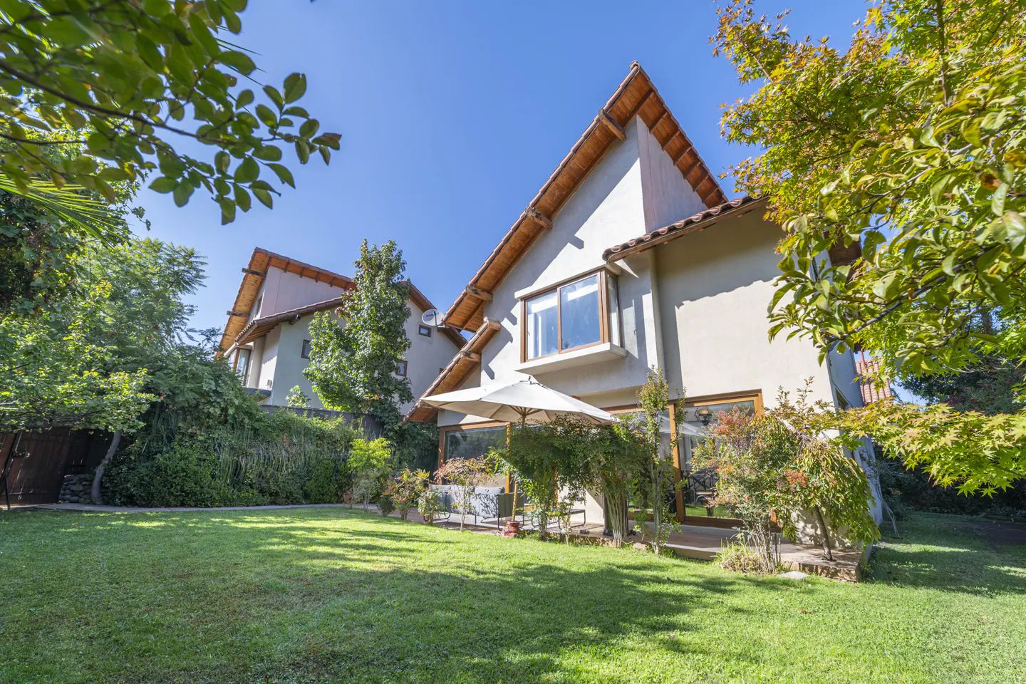 Exterior view of a two-story house with a green lawn, trees, and a white umbrella. The house has a brown roof and beige walls.