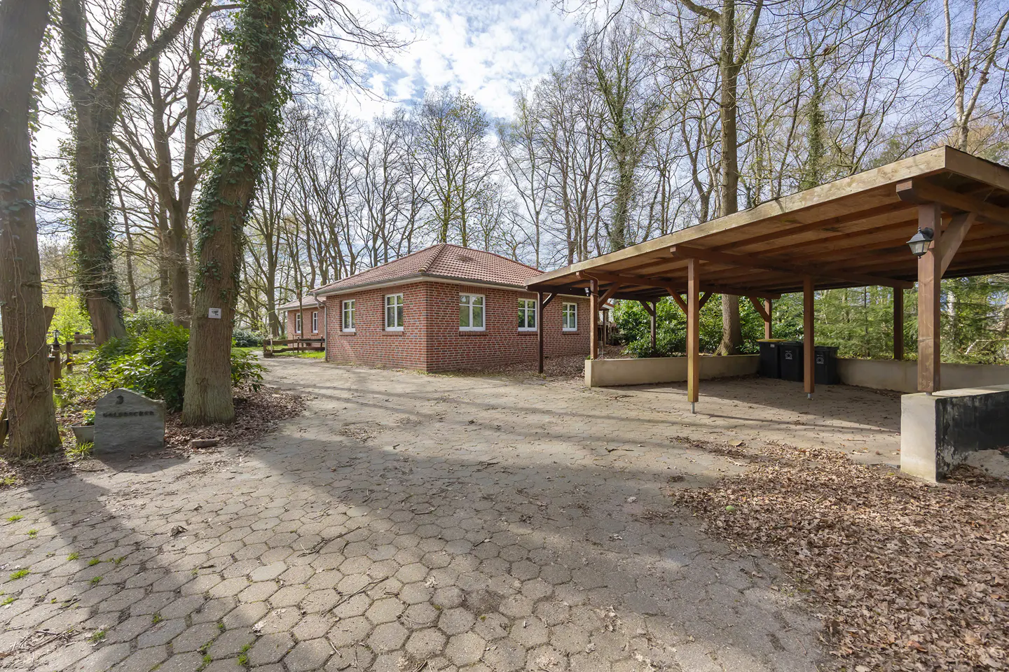 Exterior view of a red brick house with white windows, a wooden carport, and a stone driveway surrounded by trees.
