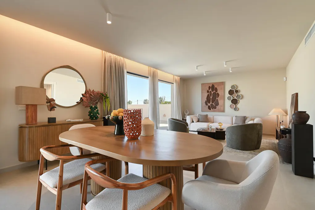 A bright, modern living and dining area with a wooden table, chairs, and a sofa in the background. Neutral tones and natural light fill the space.