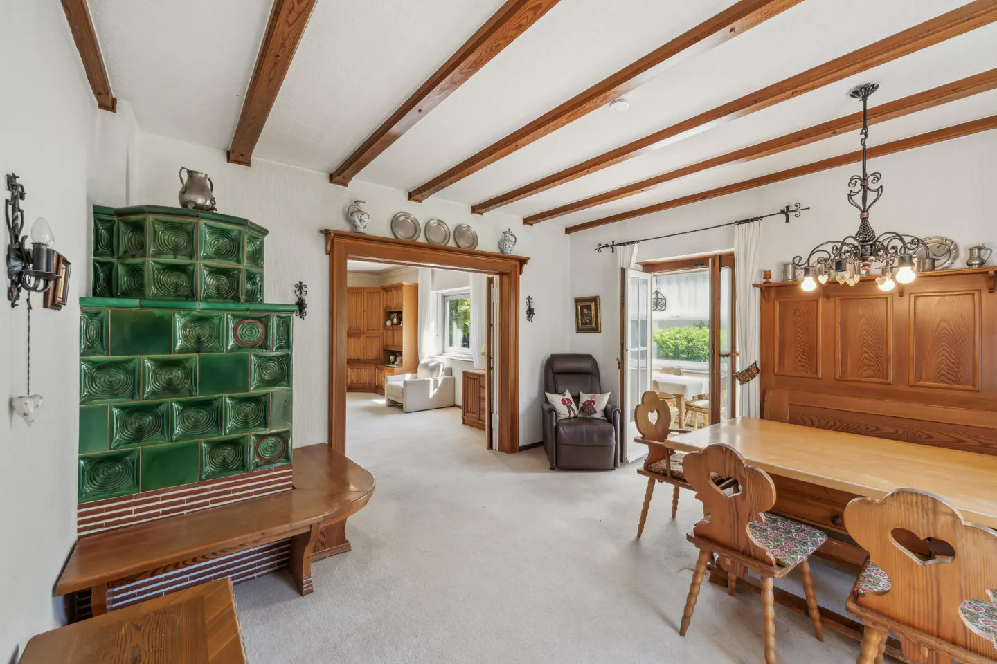 Dining room with wood-beamed ceiling, green tiled stove, wooden table with heart-cutout chairs, and view into living room.
