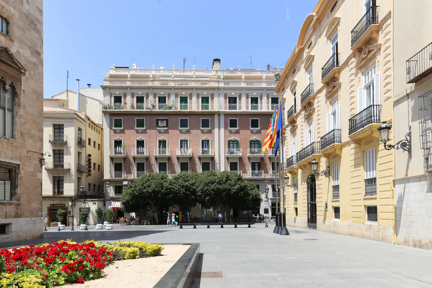 City square with red flowers, trees, and buildings. Flags are displayed in front of a yellow building with black wrought iron balconies.