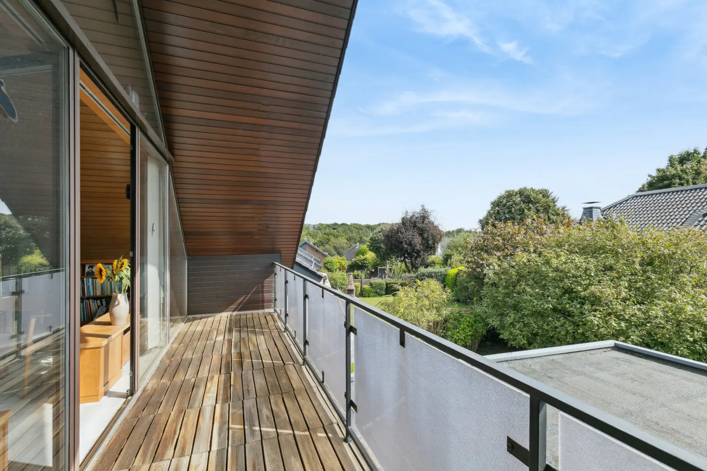 A long, narrow balcony with wood flooring and a frosted glass railing overlooks a lush green garden.
