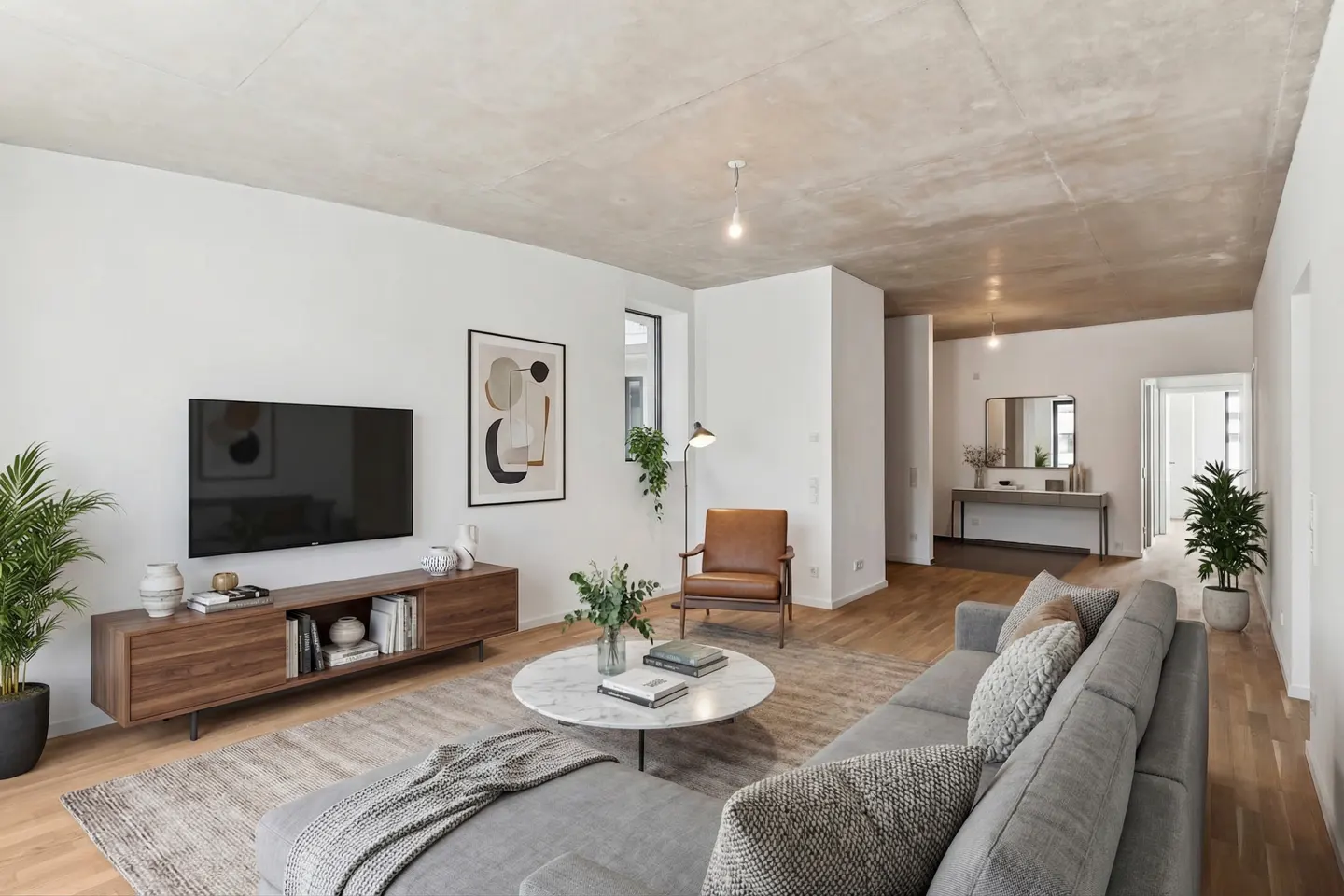 A modern living room with a gray sectional sofa, wood floors, and a concrete ceiling. A TV and artwork adorn the white walls.