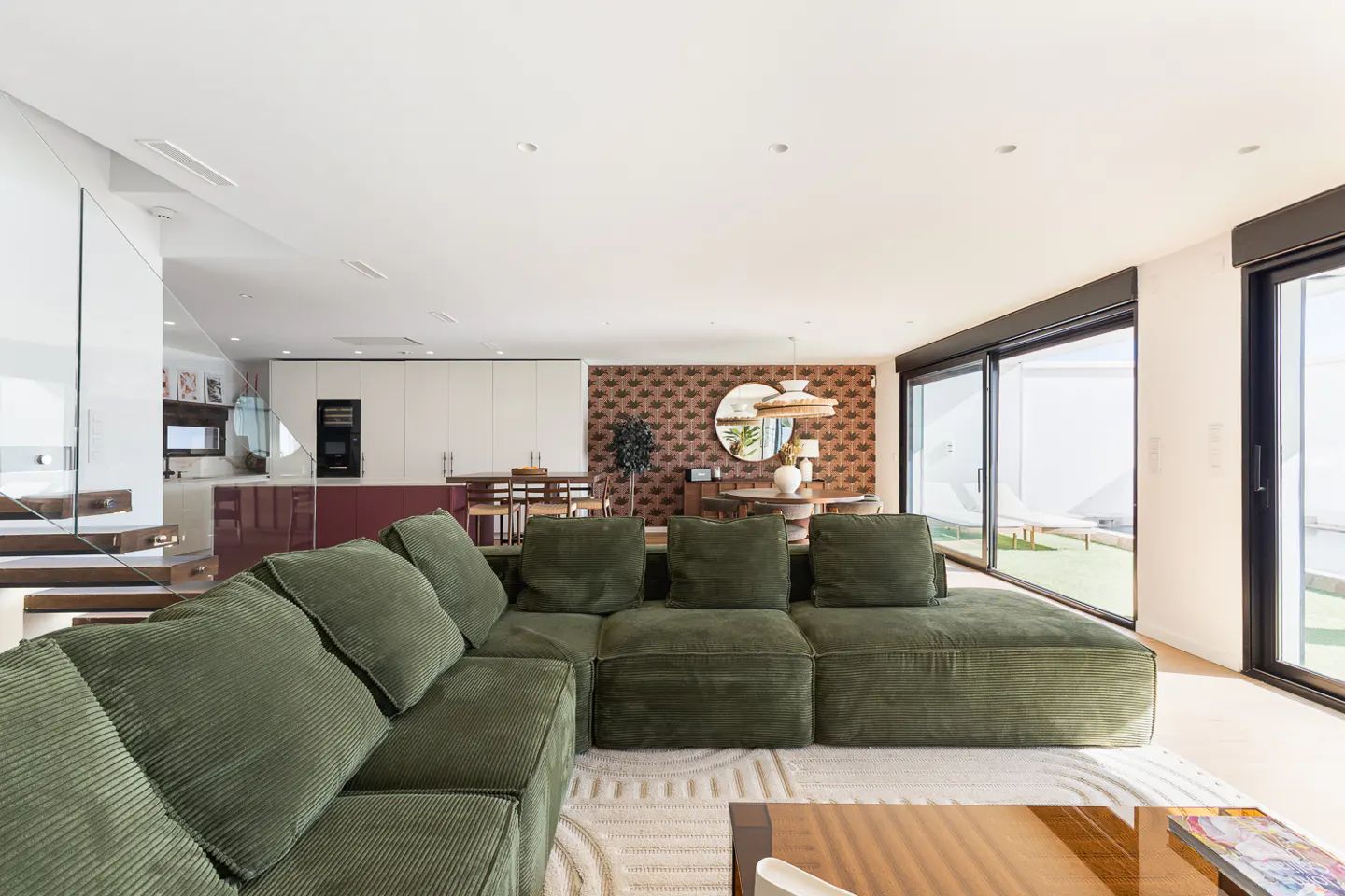 Modern open-plan living room with a large olive green corduroy sectional sofa, patterned wallpaper, and glass patio doors.