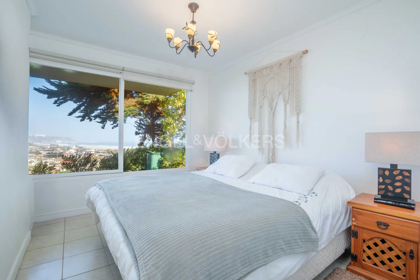 Bright bedroom with a queen bed, macrame wall hanging, chandelier, and a large window overlooking a coastal landscape.