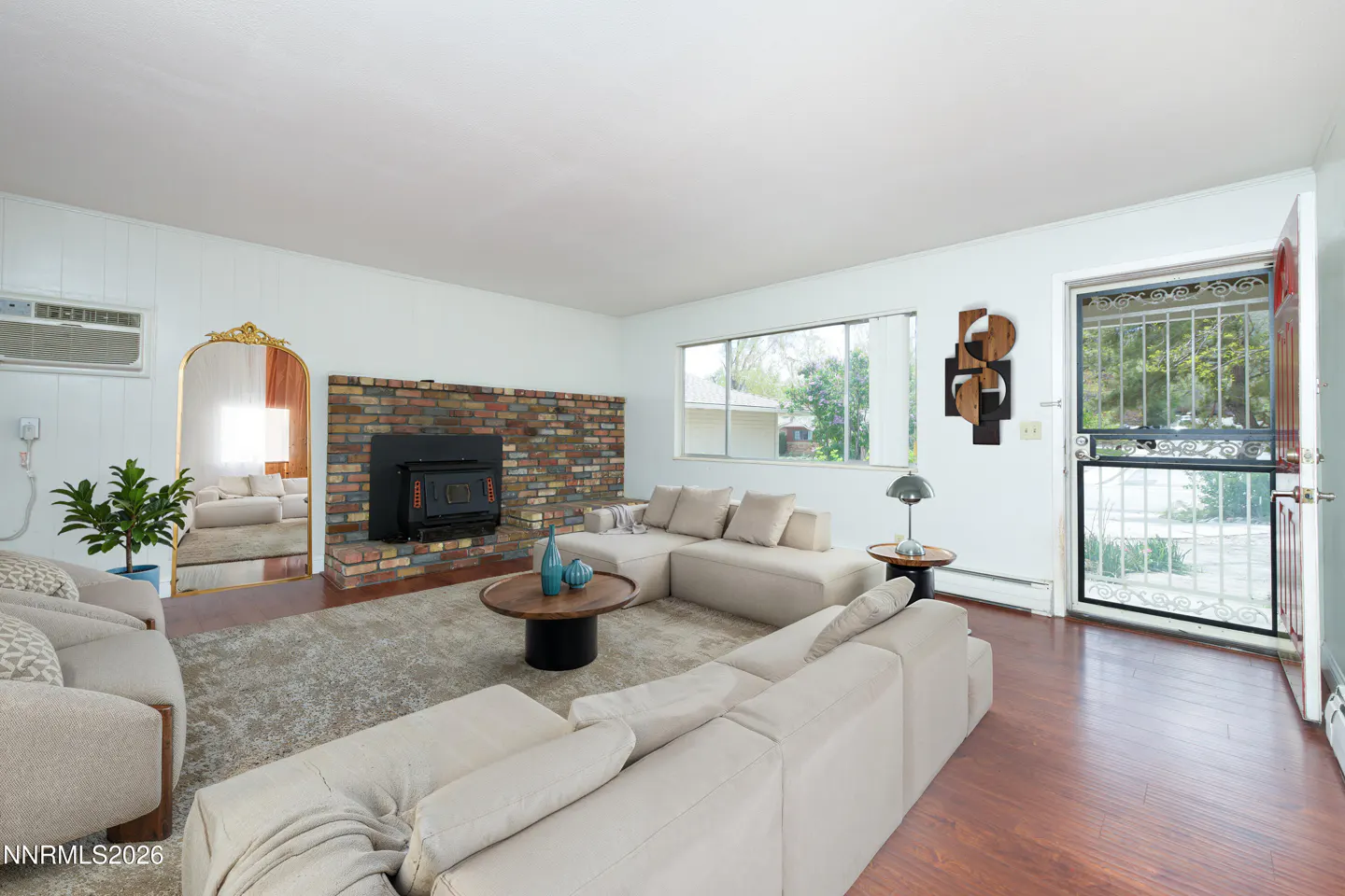 Living room with white walls, wood floors, and a brick fireplace. A beige sectional sofa sits on a gray rug. A gold mirror reflects the room.