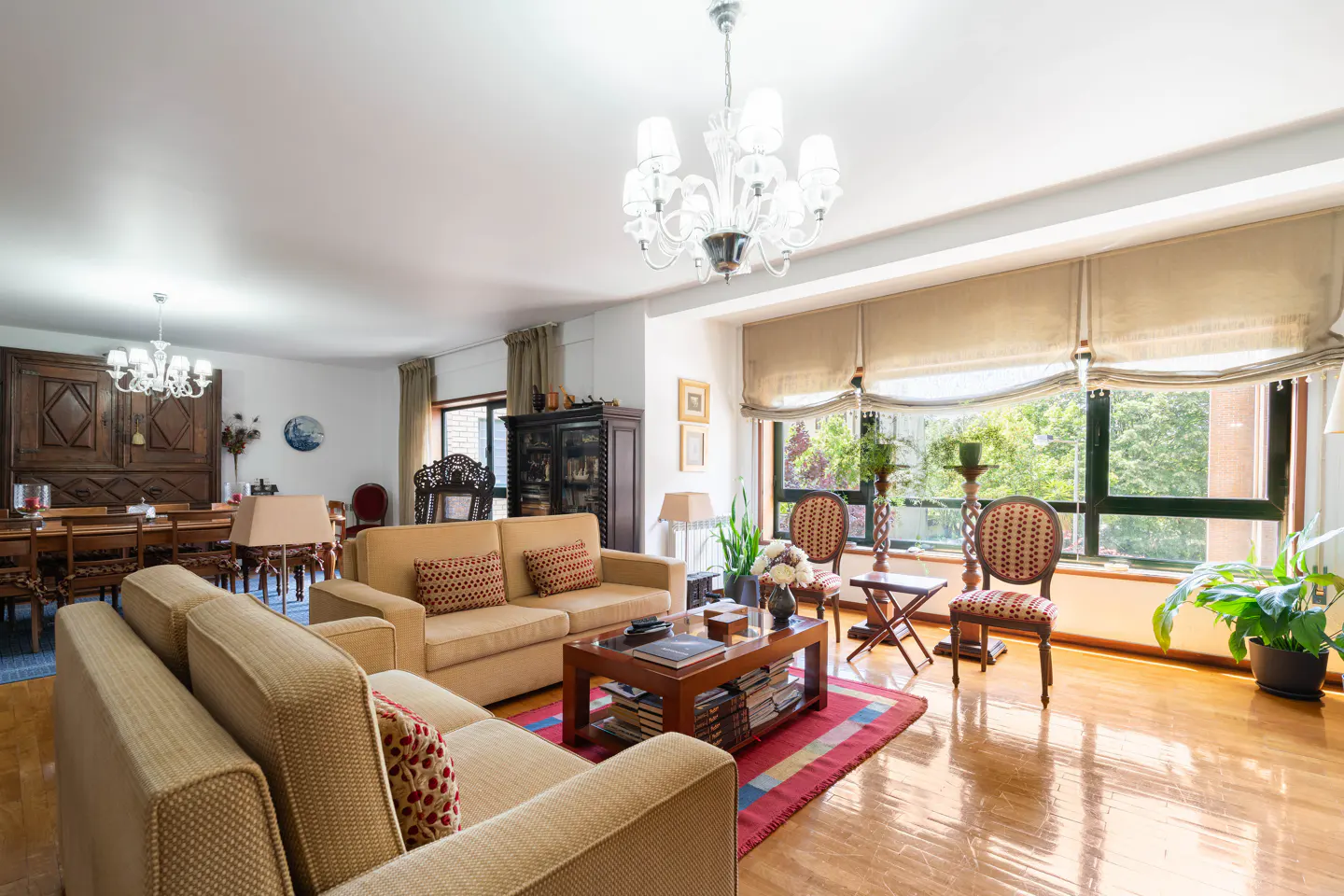 Bright living room with hardwood floors, beige sofas with patterned pillows, and a large window with a view of greenery. A chandelier hangs from the ceiling.