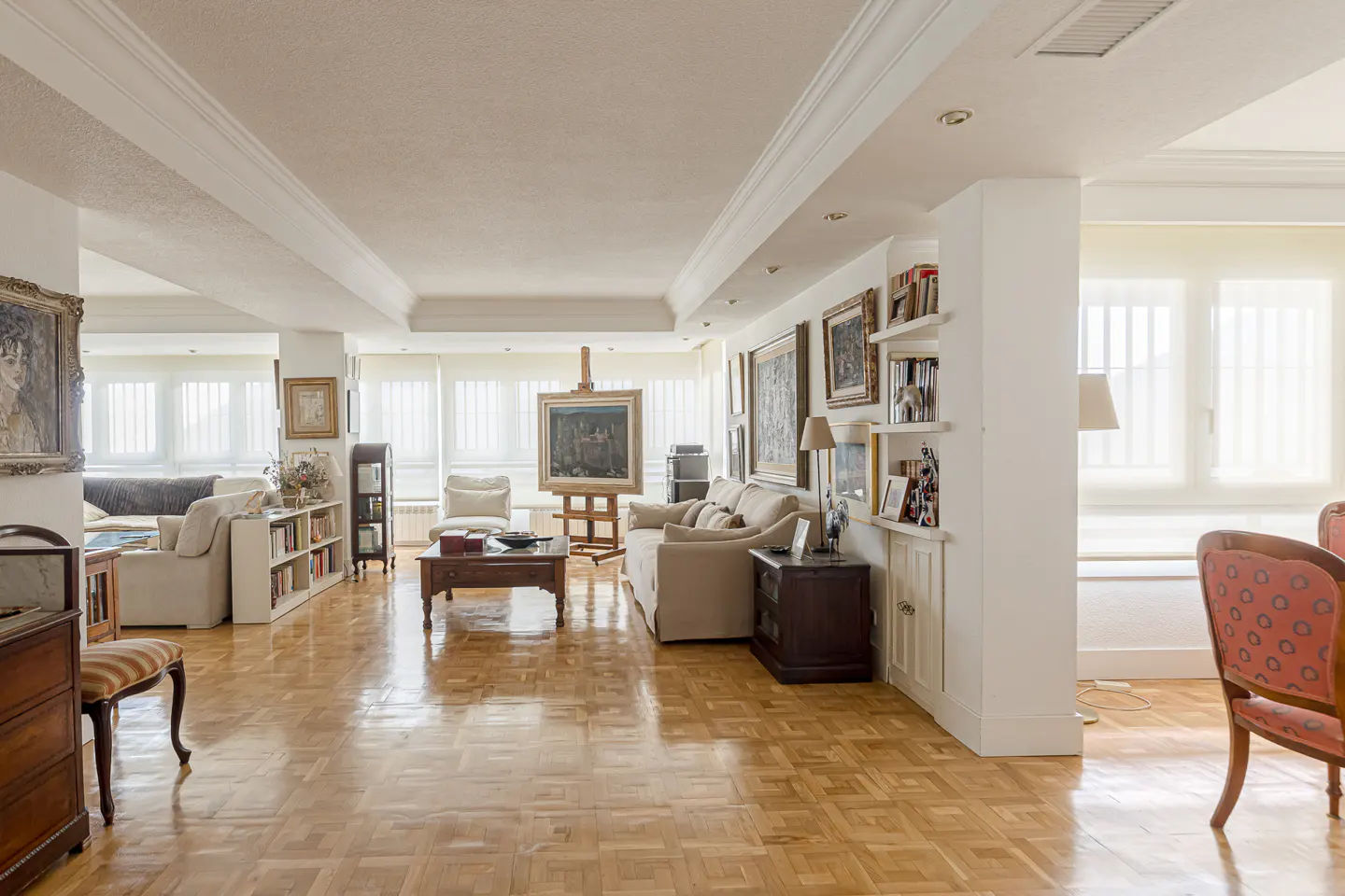 Spacious living room with parquet floors, beige sofas, an artist's easel, and walls decorated with framed paintings.