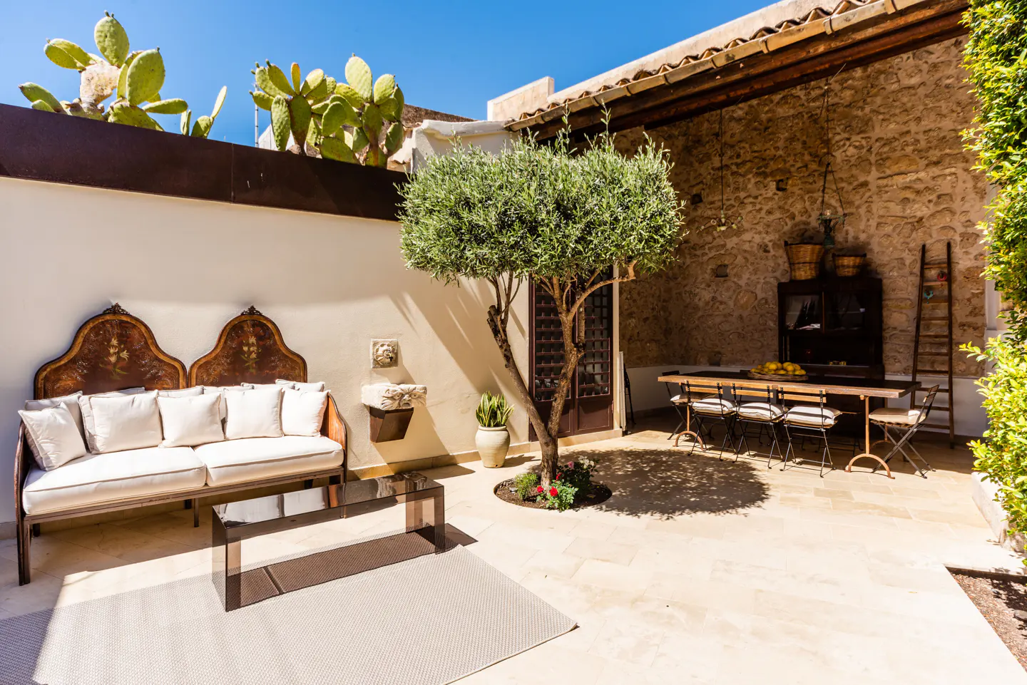 Outdoor patio with stone walls, a wooden table and chairs, and a couch with white cushions. A tree grows in the center of the patio.