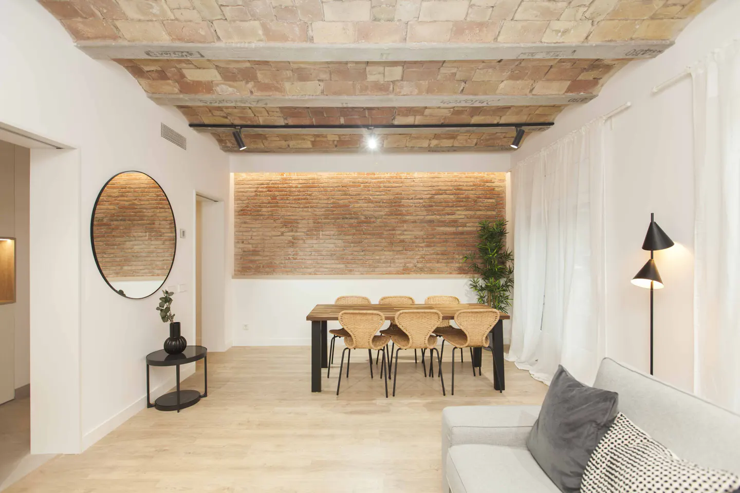 Bright living room with brick ceiling and wall. A wooden table with wicker chairs sits near a white sofa and black floor lamp.