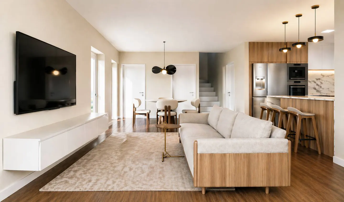 Open-concept living space with a cream sofa, dining table, and kitchen island with bar stools. A large TV is mounted on the wall.