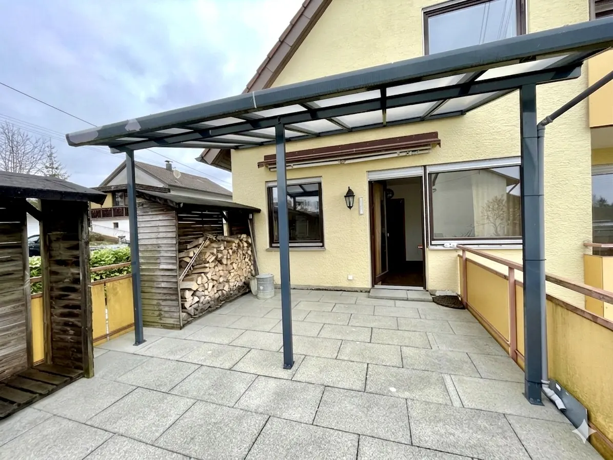 Patio with gray pavers, a metal awning, and a wood shed filled with logs next to a yellow house.