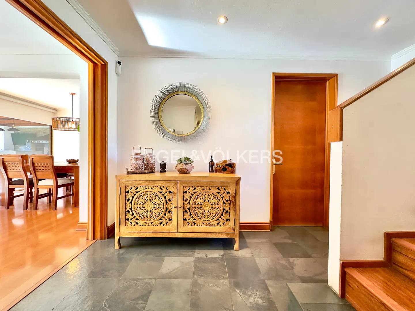 Interior shot of a home's foyer with a carved wooden cabinet, round mirror, and slate tile floor. A dining room is visible through an open doorway.