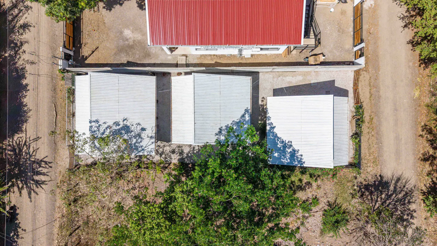 Aerial view of a property with a red-roofed house and three white-roofed outbuildings, surrounded by trees and a dirt road.
