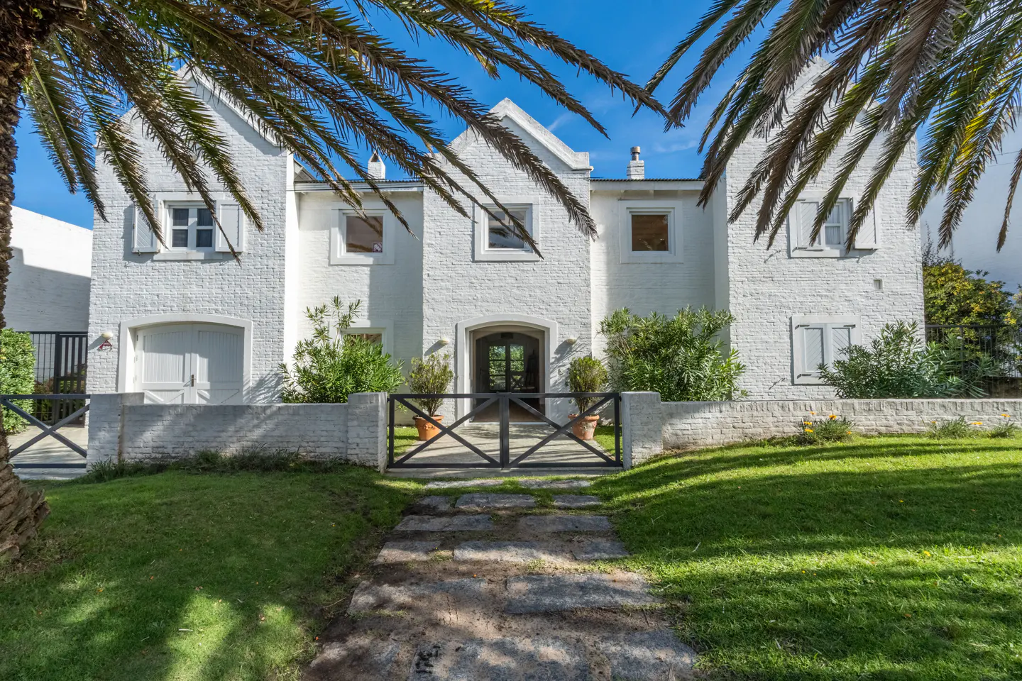 Exterior of a two-story white brick house with a black gate, green lawn, and palm trees.