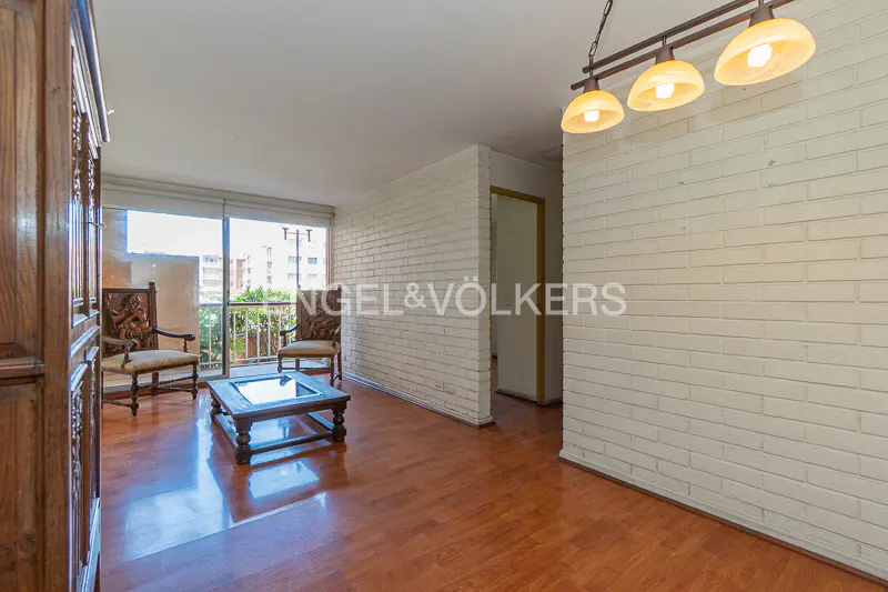 Living room with wood floors, white brick walls, and a balcony. Two carved wood chairs face a glass-topped coffee table. A chandelier hangs from the ceiling.