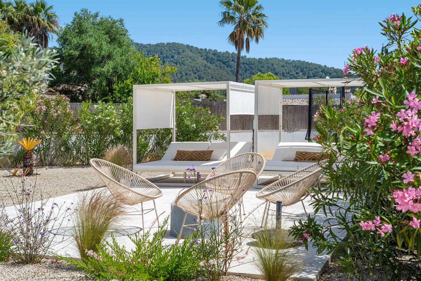 Outdoor lounge area with white canopied daybeds, woven chairs, and lush greenery under a clear blue sky.
