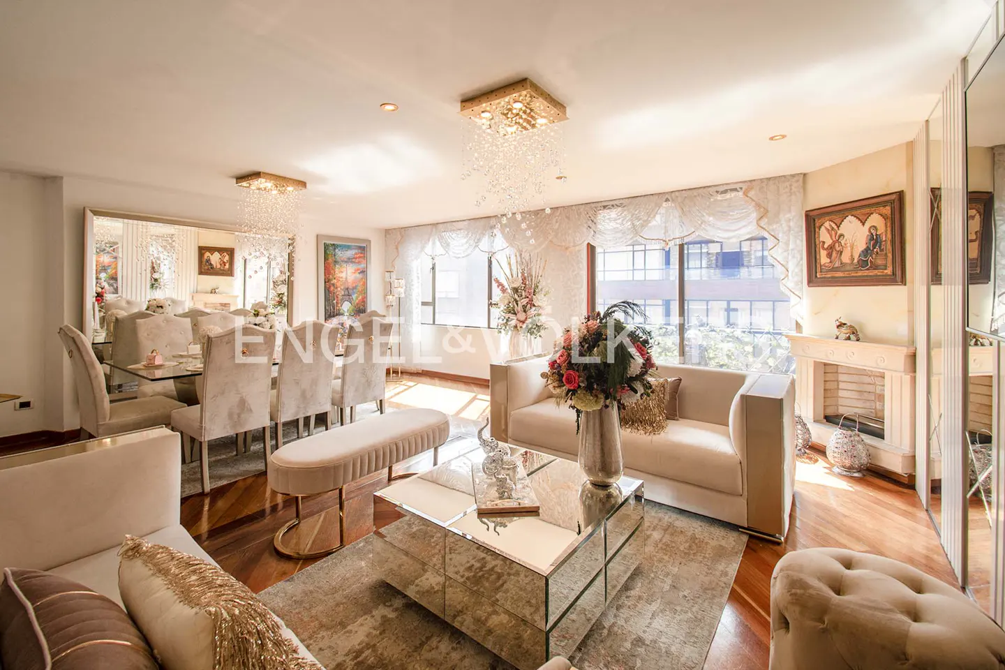 Elegant living and dining area with beige sofas, mirrored coffee table, and dining set under crystal chandeliers. Hardwood floors and large windows.