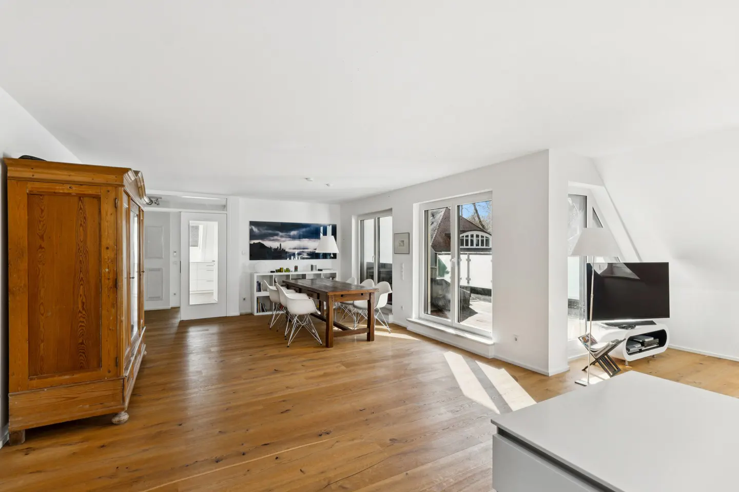 Bright, open-concept living space with wood floors, white walls, and a large wooden armoire. A dining table and chairs sit near a balcony door. A TV is visible in the background.