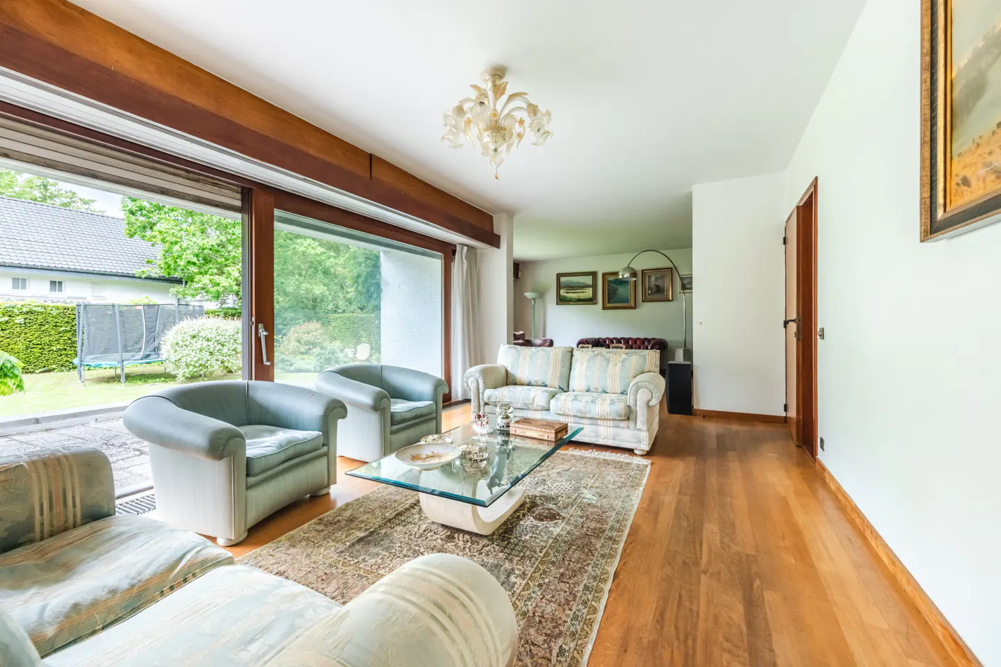 Bright living room with hardwood floors, light blue armchairs, and a patterned sofa. A glass coffee table sits on a patterned rug. Large windows overlook a green lawn.