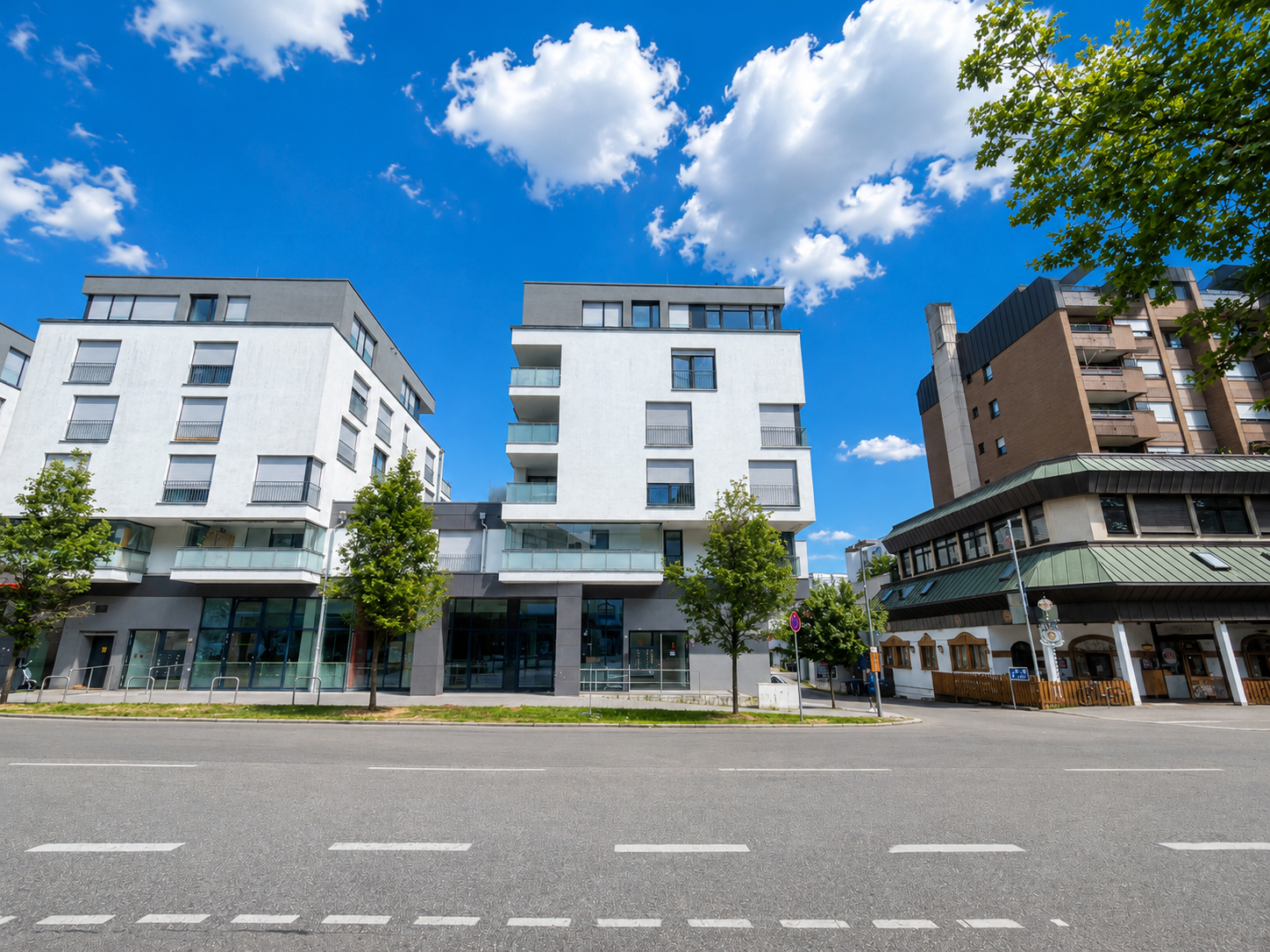 Modern white apartment buildings with balconies stand beside a traditional brown building under a bright blue sky.