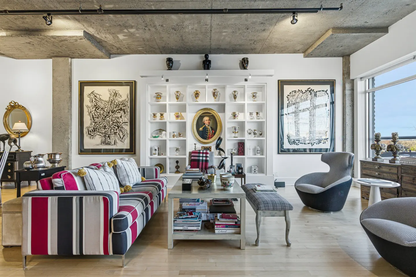 Living room with striped sofa, art, and white shelving filled with decorative objects. Concrete ceiling and light wood floors.