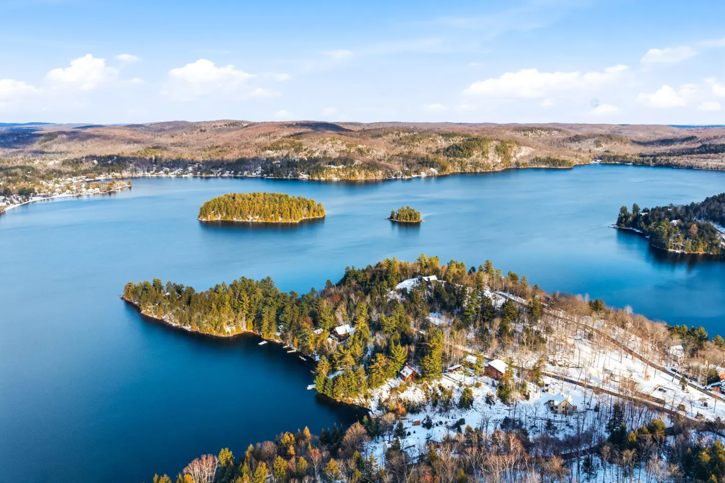 Aerial view of a blue lake with islands, surrounded by trees and some snow on a sunny day.