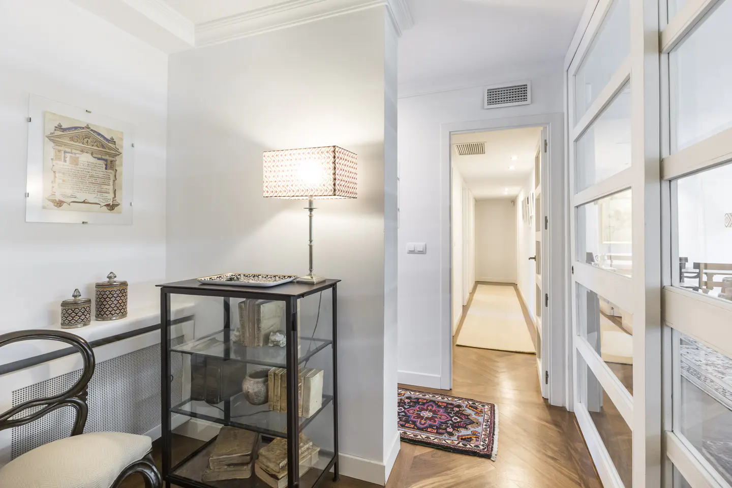 Bright hallway with a glass display cabinet, a lamp, a chair, and a view into a long corridor with hardwood floors.