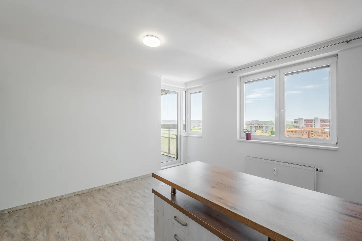 Bright, empty room with white walls, wood-look floors, and a wood-topped kitchen island. Windows overlook a city.