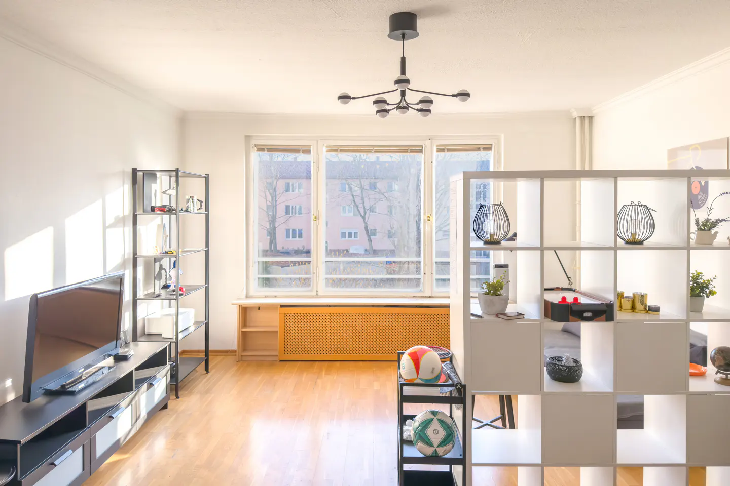 Bright living room with hardwood floors, white walls, and large window. Shelves display decorative items and sports balls. Modern chandelier hangs from the ceiling.