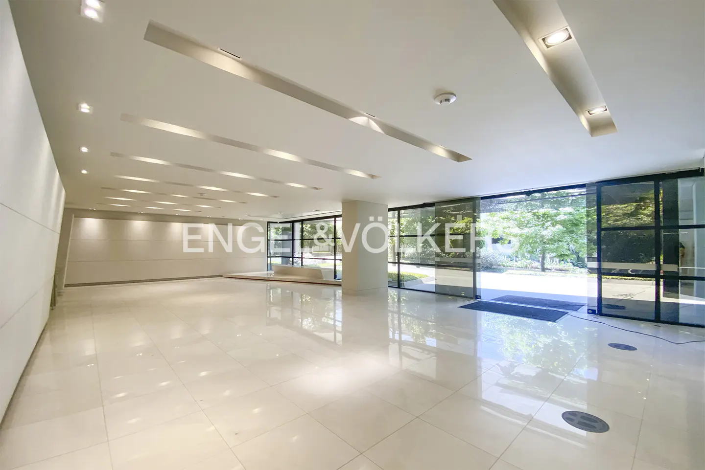 Bright, modern lobby with white tile floor and walls. Glass doors open to green trees. Engel & Völkers logo on glass.