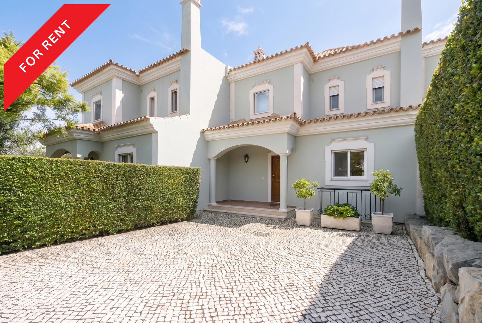 A light green two-story townhouse with a cobblestone driveway, manicured hedges, and a red "FOR RENT" banner.