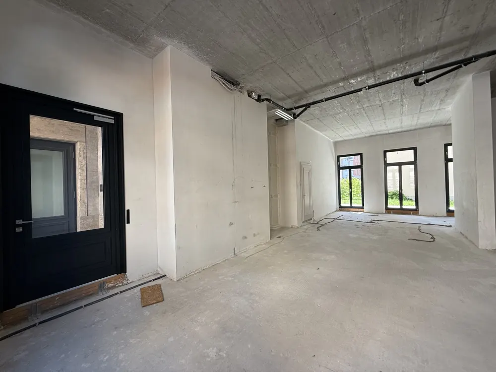 Unfinished commercial space with concrete ceiling, white walls, black door, and three large windows overlooking greenery.