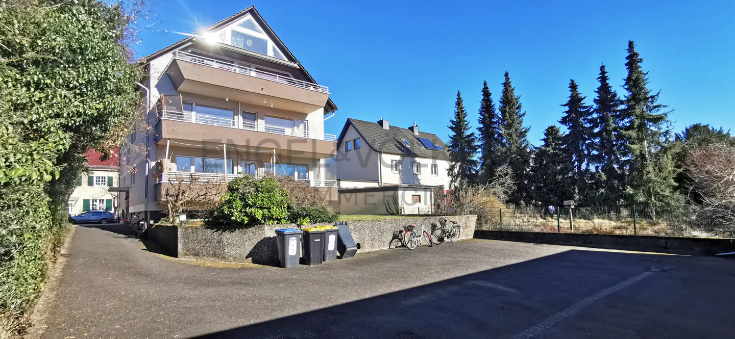 A multi-story apartment building with balconies, a paved driveway, trash bins, and tall pine trees under a clear blue sky.