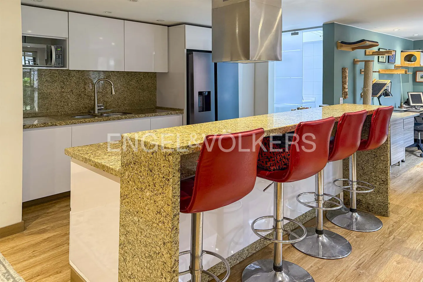 A modern kitchen with white cabinets, granite countertops, and red bar stools at the island. A stainless steel range hood is visible.