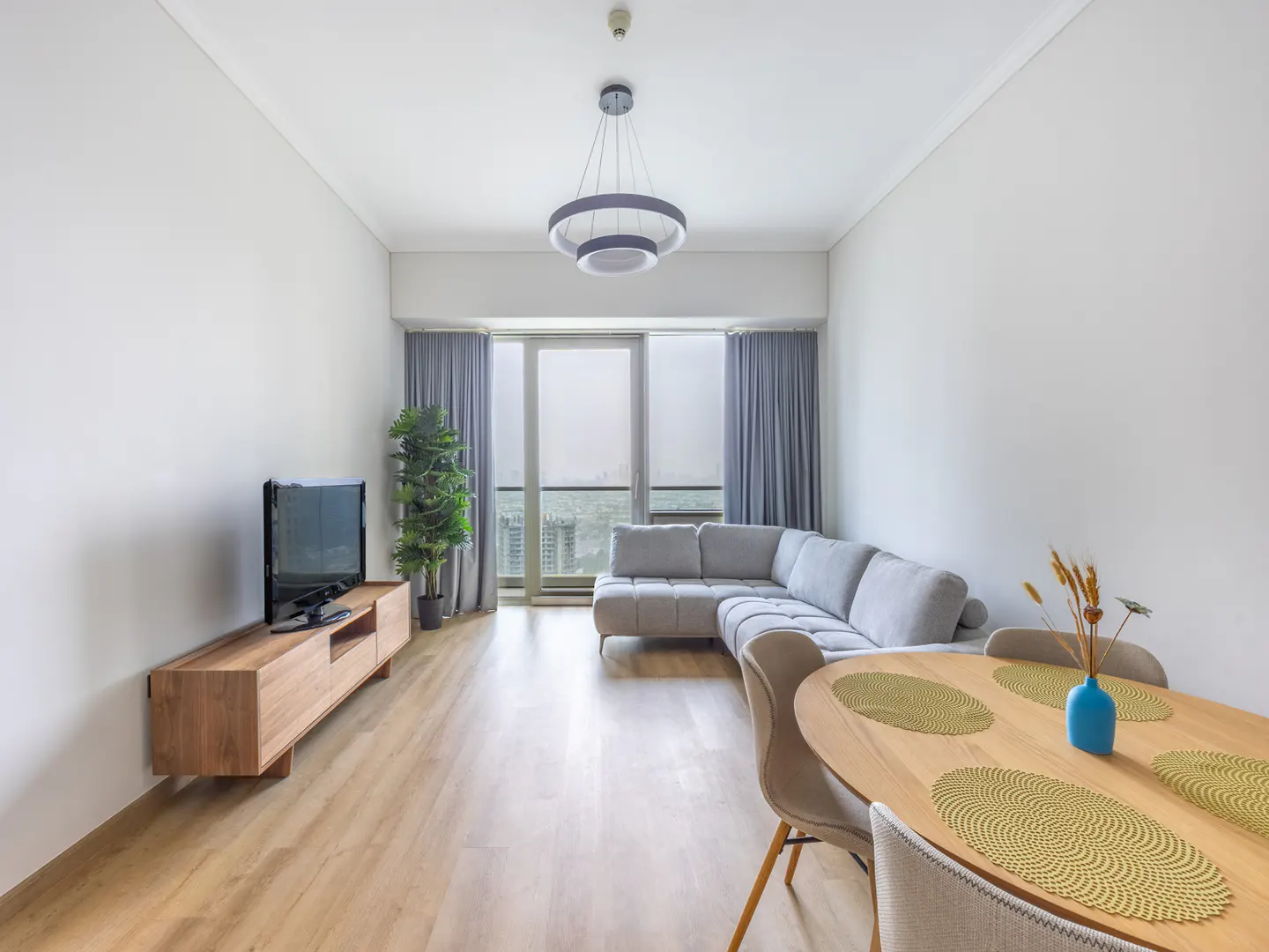 Bright living room with wood floors, white walls, and a gray sectional sofa. A wood table with place settings sits in the foreground. A TV and plant are near a window with gray curtains.