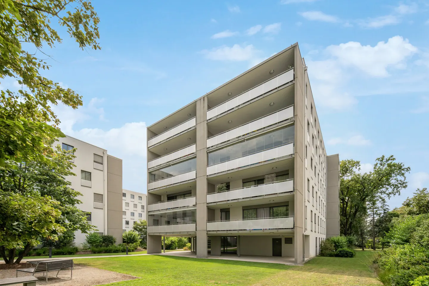 Modern apartment building with balconies, set in a green lawn with trees under a blue sky.
