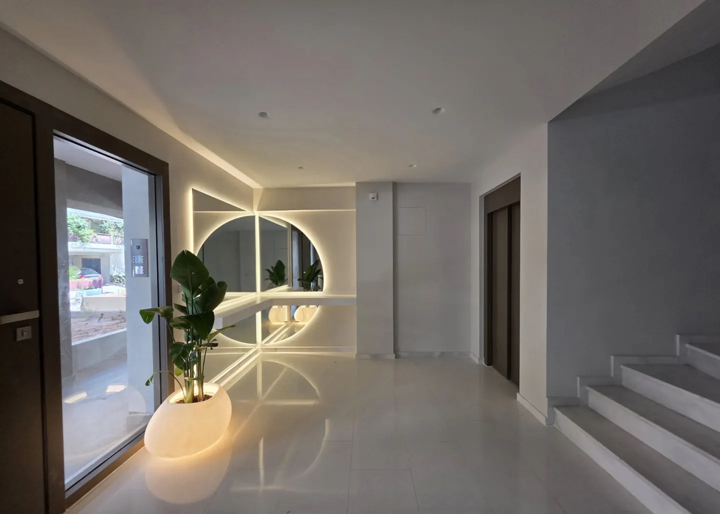 Modern building lobby with white floors, a glowing circular mirror, a potted plant, an elevator, and white stairs.