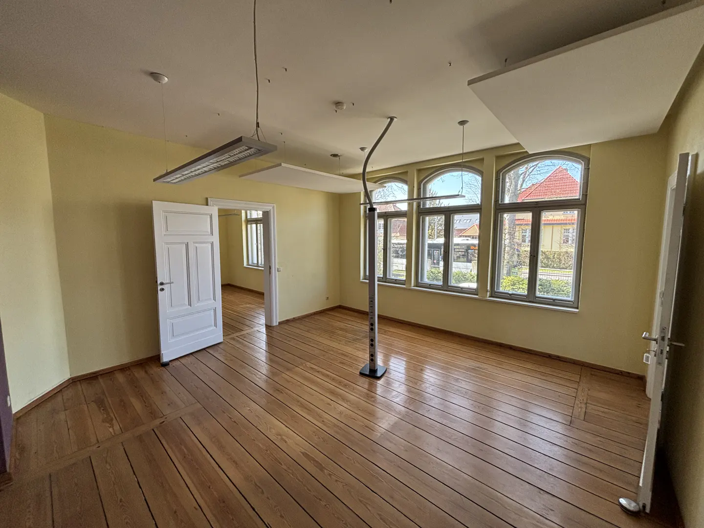 Empty room with wood floors, yellow walls, and three arched windows. A white door is open to another room. Modern lighting fixtures hang from the ceiling.