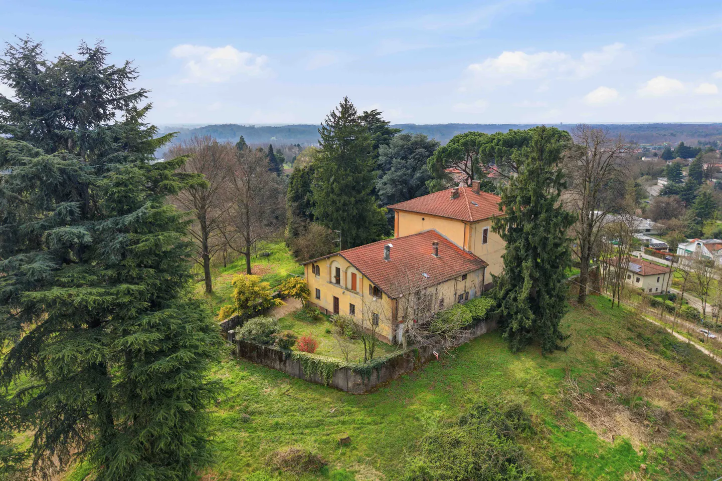 Aerial view of a yellow two-story house with a red tile roof, surrounded by green trees and grass on a hill.
