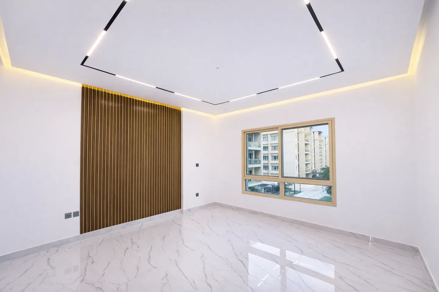 Bright, empty room with white walls, marble floors, and a window showing an apartment building. A brown wood slat wall accent and modern square lighting fixture add detail.