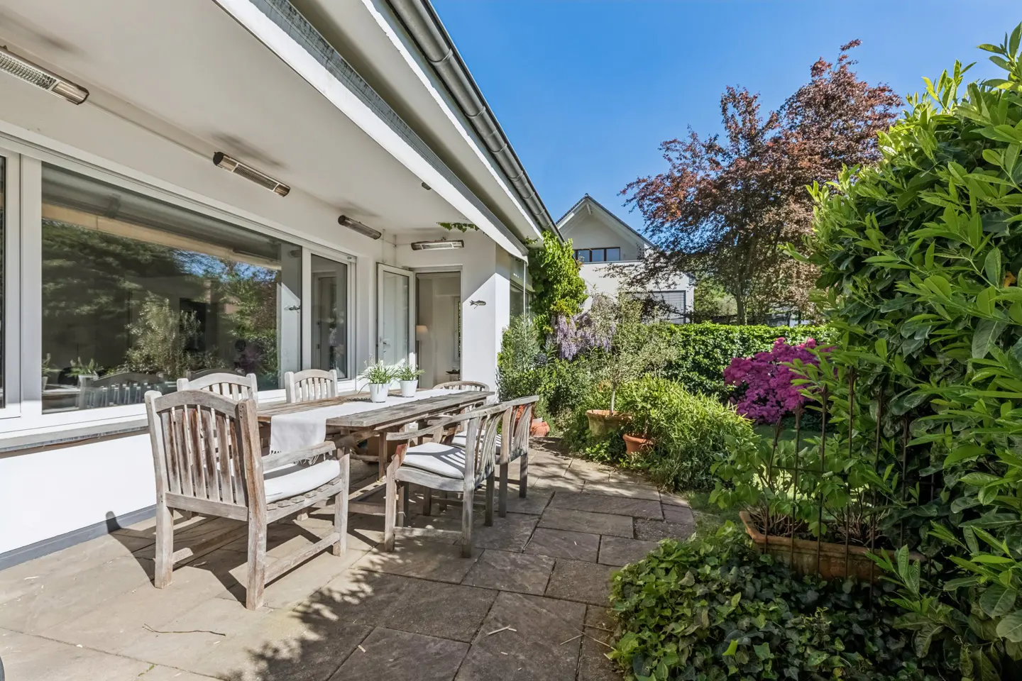 A stone patio with a wooden dining set sits outside a white house, surrounded by lush green plants and blooming flowers.