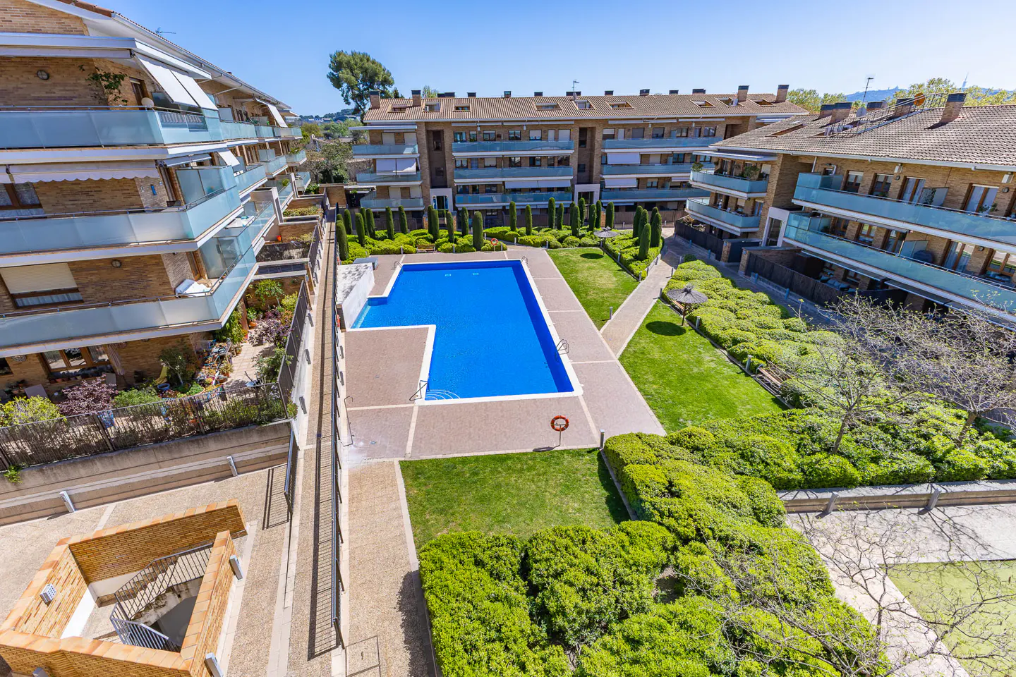 Aerial view of a bright blue swimming pool surrounded by green lawns and apartment buildings on a sunny day.
