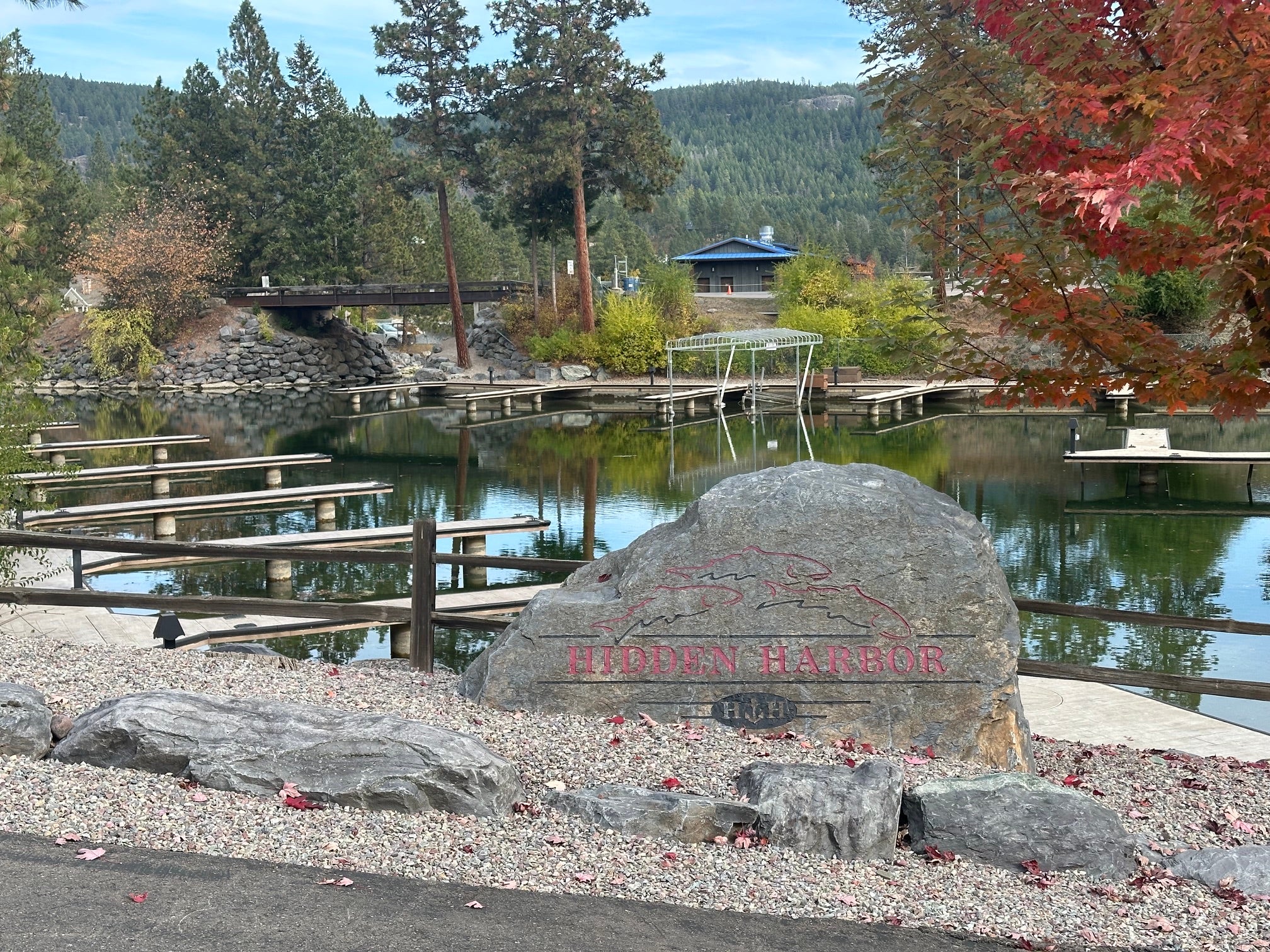 A large rock sign reading "Hidden Harbor" overlooks a calm marina with wooden docks, surrounded by trees and autumn foliage. [AI-generated text]